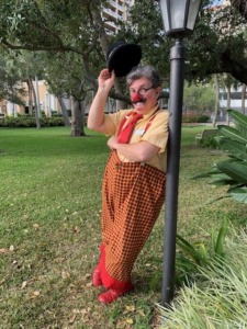 Clown performer in colorful costume and red nose, standing in park near lamp post, promoting circus arts and entertainment.
