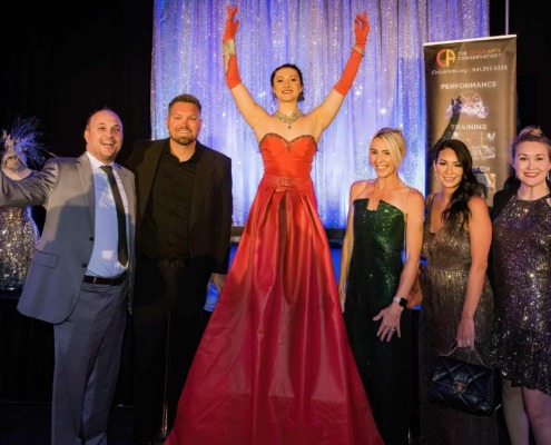 Elegant circus performer in a red gown on stage with a group of well-dressed people at a circus arts event.