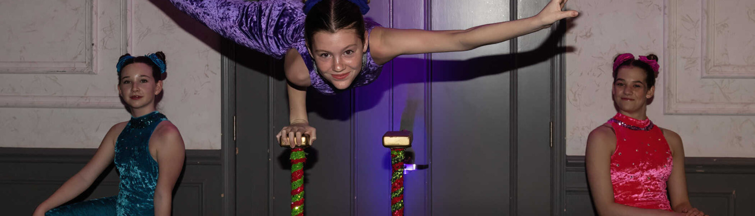 Girl performing handstand on stage with two other girls sitting nearby at circus staging event, colorful costumes, acrobatics, circus performance, youth entertainment, talent show relevance.