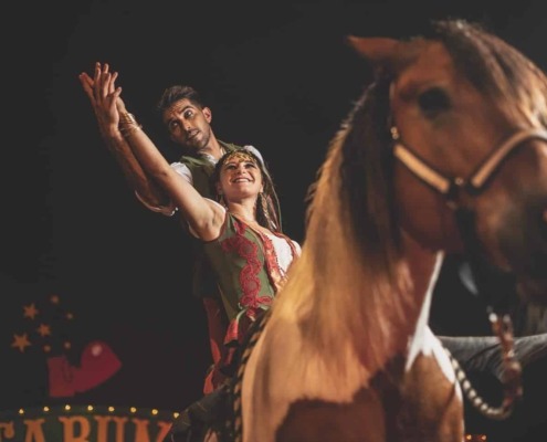 Colorful circus performers riding a brown horse with a dark background.