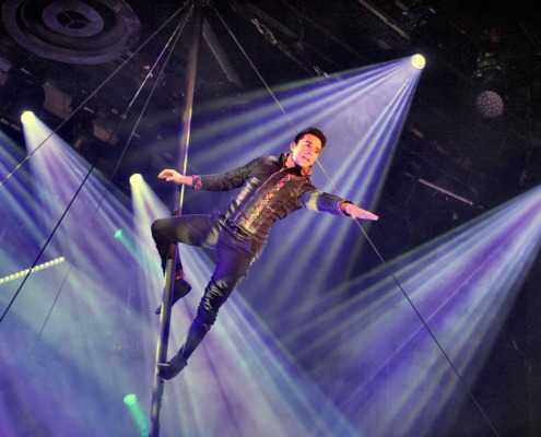Acrobat on aerial pole performing circus arts staging under vibrant stage lighting.