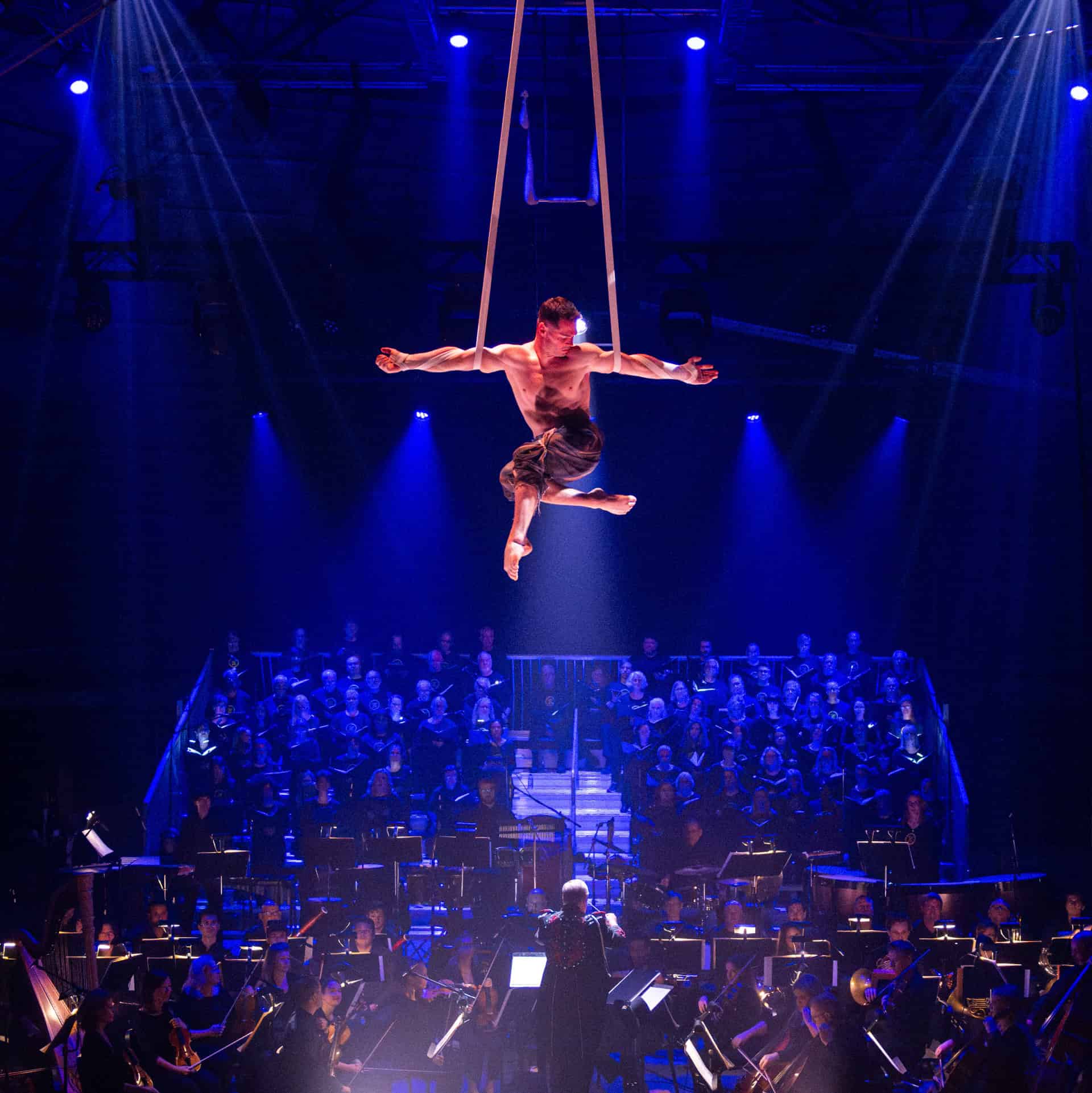 Circ-109 Dancer performing aerial act on trapeze at circus stage with orchestra and choir in background.