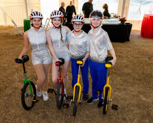 Colorful circus stagers wearing helmets and costumes holding small bikes in a circus tent setting.